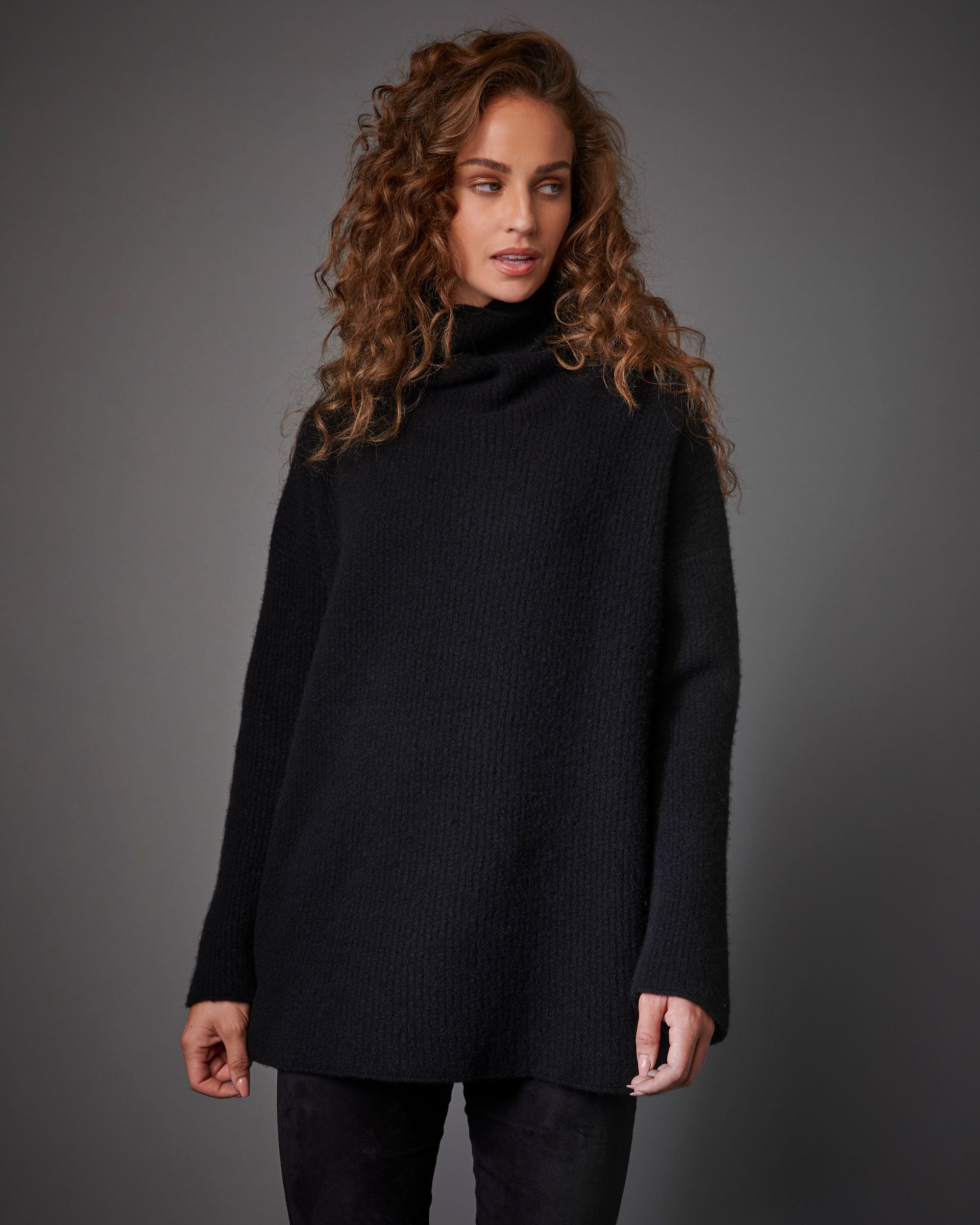 A woman wearing a long sleeve, cowl neck, black ribbed sweater with a turtleneck.